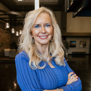 Smiling blonde woman wearing a blue ribbed sweater with arms crossed indoors.