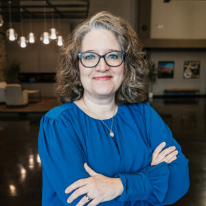 Portrait of a smiling woman with curly gray hair and blue-framed glasses, wearing a royal blue blouse, arms crossed indoors.