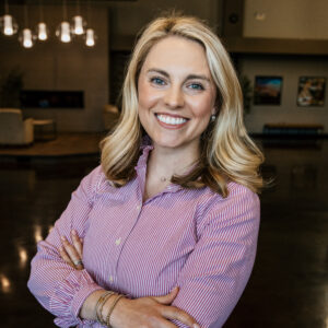 Smiling woman with blonde hair in a pink striped shirt, arms crossed indoors, looking at the camera.