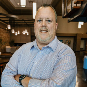 Close-up of a smiling man in a blue checkered shirt with folded arms in a cafe.
