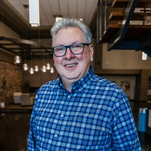Smiling man with gray hair and blue glasses wearing a blue plaid shirt indoors in an office-like space.