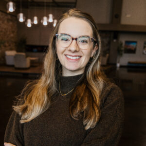 Smiling woman with tortoiseshell glasses and long light-brown hair wearing a dark brown sweater in a warmly lit indoor setting.
