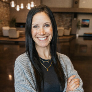 Smiling woman with long dark hair wearing a gray knitted cardigan and layered gold necklaces, indoors with warm lighting.