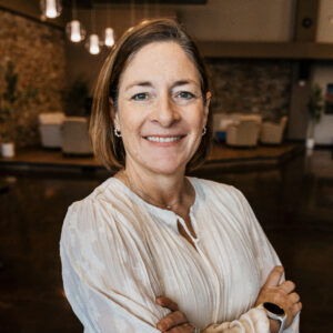 Smiling woman with light brown hair in a white textured blouse, arms crossed indoors with warm hanging lights in the background, portrait-style photo.