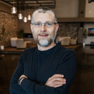 Smiling man with gray beard and glasses, arms crossed, in a warmly lit cafe setting.