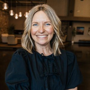 Smiling woman with blonde hair and blue eyes, wearing a dark navy blouse, indoor softly lit background.