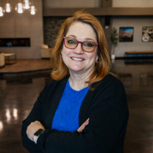 Smiling woman with red glasses and blue top, arms crossed in a warm indoor setting.