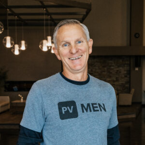 Smiling man wearing a gray shirt with a bold black rectangular graphic containing the letters MEN (indoor setting).