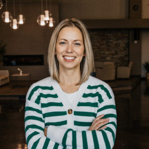 Smiling woman with blonde hair, wearing a white sweater with green stripes, arms crossed in a warmly lit room.
