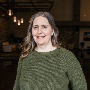 Close-up of a smiling woman wearing a green knit sweater, indoors with warm hanging lights in the background.