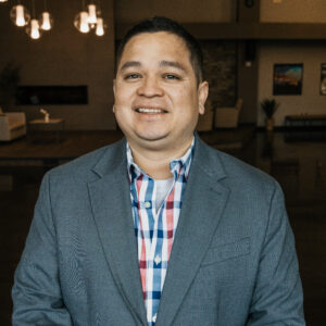 Smiling man in a blue blazer and plaid shirt in a warmly lit office background.