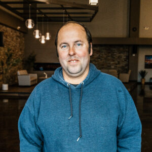 Close-up portrait of a man with a blue hoodie, indoors, with hanging pendant lights and a brick wall in the background.