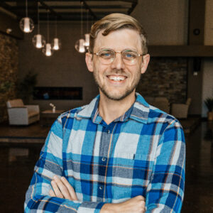 Smiling man with glasses in a blue plaid shirt, arms crossed, standing in a dimly lit cafe.