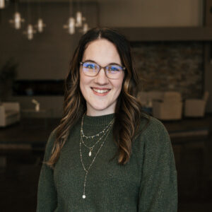Close-up portrait of a smiling woman with long brown hair, wearing glasses, a dark green sweater, and layered silver necklaces indoors.