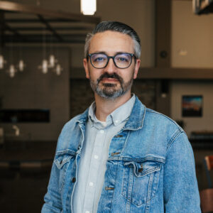 Man with gray beard and glasses wearing a denim jacket, indoors.