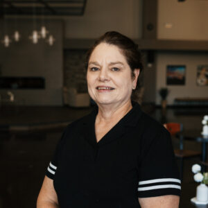 Portrait of a woman with dark hair wearing a black top with white stripes on the sleeves, smiling at the camera in a blurred indoor setting.