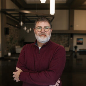 Man with gray beard and round glasses wearing a maroon quarter-zip sweater, arms crossed indoors.
