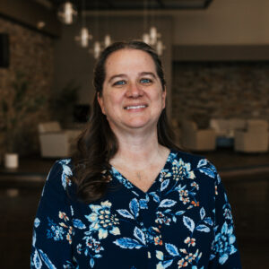 Smiling woman wearing a navy blue floral blouse, standing indoors with a blurred background.