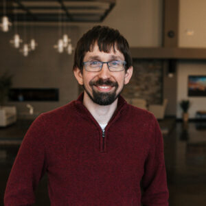 Smiling man with dark hair and beard wearing glasses and a maroon knit sweater indoors, background blurred.