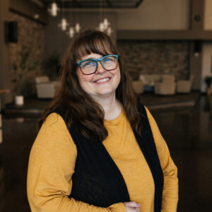 Portrait of a smiling woman with teal-framed glasses wearing a mustard yellow top and dark overalls, standing indoors with a blurred background.