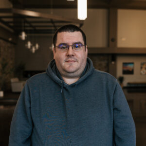 Portrait of a man with short dark hair, wearing glasses and a blue hoodie, indoors.