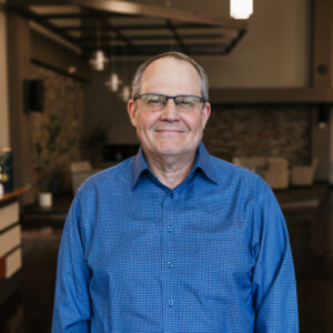Smiling man wearing a blue checkered shirt and glasses indoors, facing the camera.